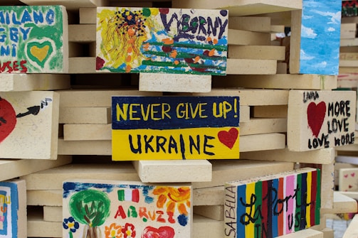 Several painted bricks are stacked together, each featuring colorful artwork and positive messages. The central brick says 'Never give up! Ukraine' with a red heart, while others display abstract art and phrases promoting love and unity.