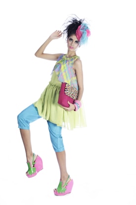 A fashion-forward individual poses in a vibrant and eclectic outfit. The ensemble includes a pastel plaid top, a lime green dress, and light blue capri pants. Accessories include a large pink and blue bow in the hair, a bright pink clutch, and matching pink wedge sandals with green accents. The hair is styled in a voluminous updo, and the person exudes a playful and quirky charm.
