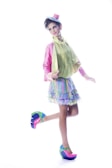 A person is dressed in vibrant, colorful clothing, including a layered skirt with pastel hues, a lime green blouse, and a pink jacket. The outfit is accessorized with a small pink top hat featuring a blue ribbon. The footwear is a pair of platform shoes with a bright, multicolored design. The individual poses with one foot raised, exuding a playful and whimsical style.