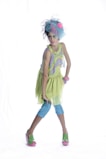 A young girl is dressed in a colorful, whimsical outfit, consisting of a light green dress with a ruffled skirt, blue leggings, and green sandals with pink soles. She is accessorized with a large blue headpiece with a pink flower, and multiple pink bracelets on her wrist. She strikes a confident and poised pose against a plain white background.