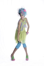A young girl is dressed in a colorful, whimsical outfit, consisting of a light green dress with a ruffled skirt, blue leggings, and green sandals with pink soles. She is accessorized with a large blue headpiece with a pink flower, and multiple pink bracelets on her wrist. She strikes a confident and poised pose against a plain white background.
