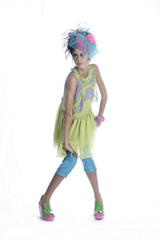 A young girl is dressed in a colorful, whimsical outfit, consisting of a light green dress with a ruffled skirt, blue leggings, and green sandals with pink soles. She is accessorized with a large blue headpiece with a pink flower, and multiple pink bracelets on her wrist. She strikes a confident and poised pose against a plain white background.