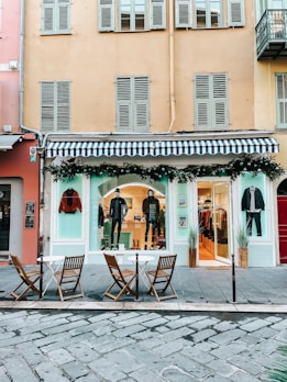 A fashion boutique with a blue and white striped awning on the ground floor of a building. Mannequins in stylish attire are displayed in the window, and there are decorations with greenery and string lights. In front of the store, there are three wooden chairs and two small round tables on a cobblestone pavement.