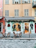 A fashion boutique with a blue and white striped awning on the ground floor of a building. Mannequins in stylish attire are displayed in the window, and there are decorations with greenery and string lights. In front of the store, there are three wooden chairs and two small round tables on a cobblestone pavement.