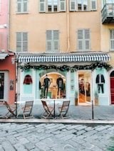 A fashion boutique with a blue and white striped awning on the ground floor of a building. Mannequins in stylish attire are displayed in the window, and there are decorations with greenery and string lights. In front of the store, there are three wooden chairs and two small round tables on a cobblestone pavement.