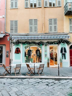 A fashion boutique with a blue and white striped awning on the ground floor of a building. Mannequins in stylish attire are displayed in the window, and there are decorations with greenery and string lights. In front of the store, there are three wooden chairs and two small round tables on a cobblestone pavement.
