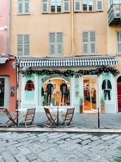 A fashion boutique with a blue and white striped awning on the ground floor of a building. Mannequins in stylish attire are displayed in the window, and there are decorations with greenery and string lights. In front of the store, there are three wooden chairs and two small round tables on a cobblestone pavement.