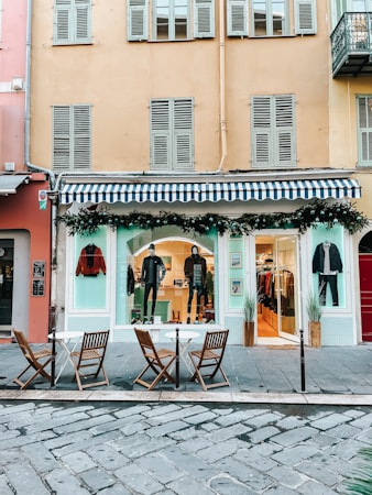 A fashion boutique with a blue and white striped awning on the ground floor of a building. Mannequins in stylish attire are displayed in the window, and there are decorations with greenery and string lights. In front of the store, there are three wooden chairs and two small round tables on a cobblestone pavement.