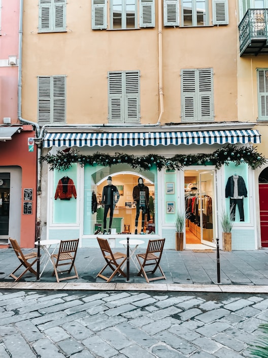 A fashion boutique with a blue and white striped awning on the ground floor of a building. Mannequins in stylish attire are displayed in the window, and there are decorations with greenery and string lights. In front of the store, there are three wooden chairs and two small round tables on a cobblestone pavement.