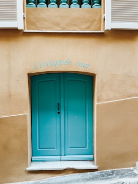 A turquoise-colored door with a heart-shaped keyhole set in a beige wall, flanked by white shutters and a railing with turquoise balusters above. The text above the door reads 'L'&eacute;chapp&eacute;e mer.'