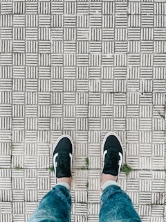 A pair of feet wearing black sneakers with white soles and stripes, positioned on a patterned, textured sidewalk. The surface consists of repeating rectangular tiles with grooves, some of which have small patches of grass growing between them. Blue jeans are also visible.