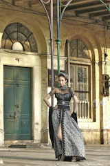 A woman dressed in a flowing Victorian black lace gown standing against a shadowy, ornate backdrop.