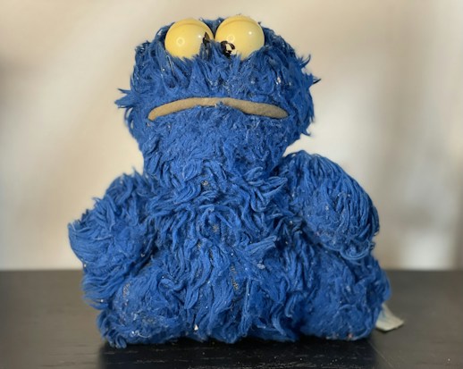 A plush toy with shaggy blue fur and large yellow eyes sits on a dark surface. The toy has a simple, cartoonish appearance with a neutral expression.