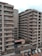 A modern apartment complex with multiple stories and balconies, featuring large windows and a neutral color palette. The surroundings include a paved entrance and a small garden area with plants and decorative elements. A few parked cars are visible in designated spaces in the foreground.