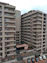 A modern apartment complex with multiple stories and balconies, featuring large windows and a neutral color palette. The surroundings include a paved entrance and a small garden area with plants and decorative elements. A few parked cars are visible in designated spaces in the foreground.