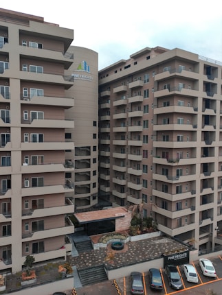 A modern apartment complex with multiple stories and balconies, featuring large windows and a neutral color palette. The surroundings include a paved entrance and a small garden area with plants and decorative elements. A few parked cars are visible in designated spaces in the foreground.