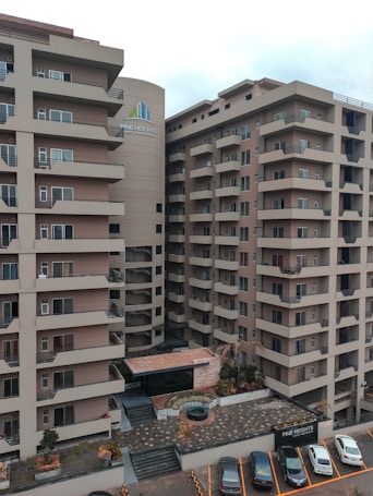 A modern apartment complex with multiple stories and balconies, featuring large windows and a neutral color palette. The surroundings include a paved entrance and a small garden area with plants and decorative elements. A few parked cars are visible in designated spaces in the foreground.