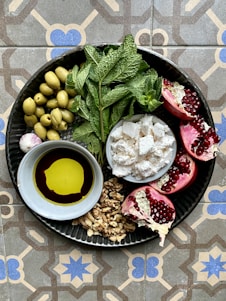 A round metal plate contains a variety of fresh and vibrant ingredients. There are green olives, fresh mint leaves, pomegranate halves with bright red seeds, crumbled cheese, a small bowl of olive oil and balsamic vinegar, and a cluster of walnuts. The plate is placed on a decorative, geometric patterned tile surface.