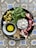 A round metal plate contains a variety of fresh and vibrant ingredients. There are green olives, fresh mint leaves, pomegranate halves with bright red seeds, crumbled cheese, a small bowl of olive oil and balsamic vinegar, and a cluster of walnuts. The plate is placed on a decorative, geometric patterned tile surface.