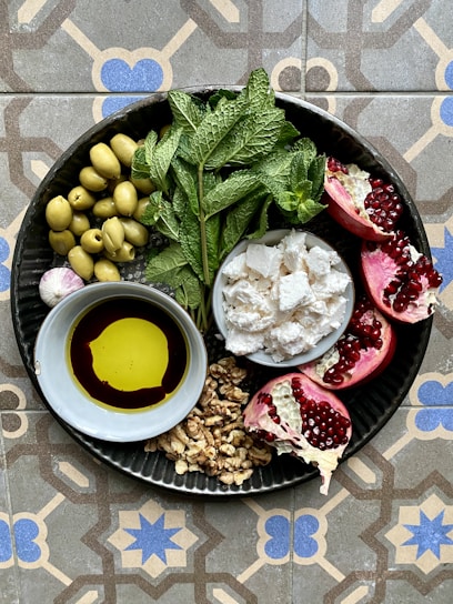 A round metal plate contains a variety of fresh and vibrant ingredients. There are green olives, fresh mint leaves, pomegranate halves with bright red seeds, crumbled cheese, a small bowl of olive oil and balsamic vinegar, and a cluster of walnuts. The plate is placed on a decorative, geometric patterned tile surface.