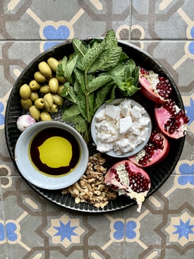 A round metal plate contains a variety of fresh and vibrant ingredients. There are green olives, fresh mint leaves, pomegranate halves with bright red seeds, crumbled cheese, a small bowl of olive oil and balsamic vinegar, and a cluster of walnuts. The plate is placed on a decorative, geometric patterned tile surface.