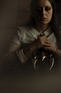 A person with long dark hair sits in a dimly lit environment. They are wearing a light-colored, long-sleeved top and are holding an ornate dagger with both hands. The facial expression is serious, and the overall atmosphere is somber and contemplative.