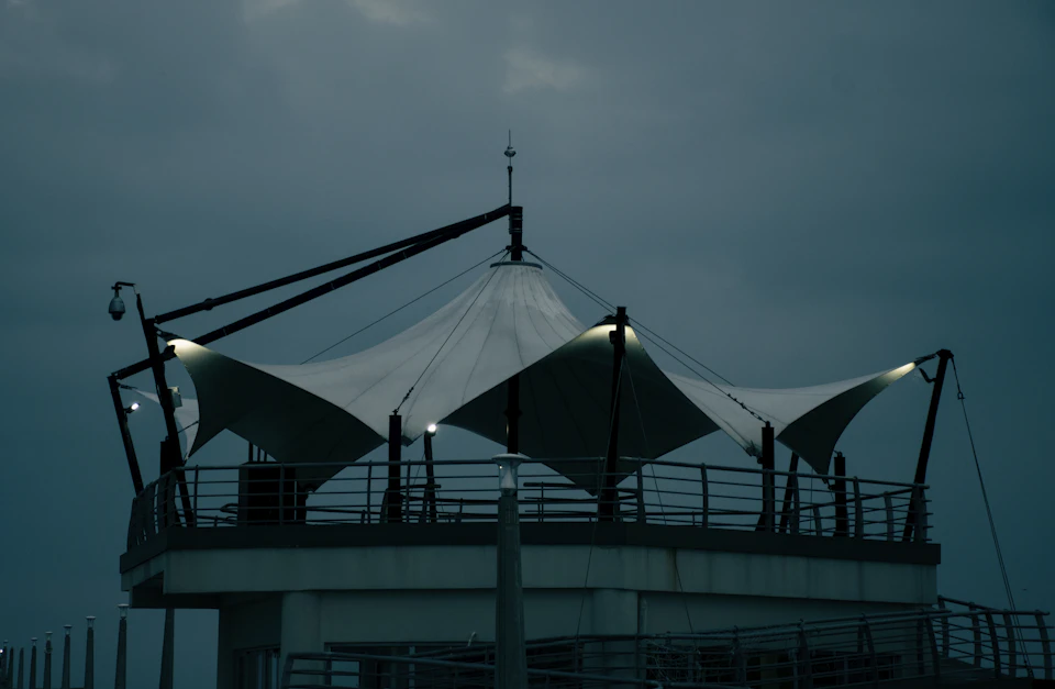 The image depicts a large white tent with three triangular sections, each adorned with small lights on top, situated atop a gray building. The tent is supported by two sturdy black poles and features a pointed roof that adds to its unique appearance. The scene is set against a dark blue sky, creating an atmospheric backdrop for the structure.