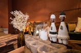 The image depicts a cozy living space with warm lighting. On a wooden table, there are decorative objects including two intricately woven African figurines, a vase with dried flowers, and a book titled about West African architecture. The background features a comfortable seating arrangement with patterned cushions.