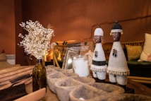 The image depicts a cozy living space with warm lighting. On a wooden table, there are decorative objects including two intricately woven African figurines, a vase with dried flowers, and a book titled about West African architecture. The background features a comfortable seating arrangement with patterned cushions.