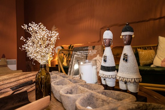 The image depicts a cozy living space with warm lighting. On a wooden table, there are decorative objects including two intricately woven African figurines, a vase with dried flowers, and a book titled about West African architecture. The background features a comfortable seating arrangement with patterned cushions.