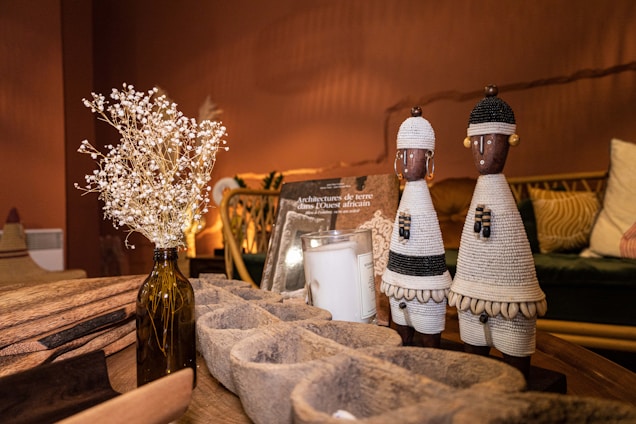 The image depicts a cozy living space with warm lighting. On a wooden table, there are decorative objects including two intricately woven African figurines, a vase with dried flowers, and a book titled about West African architecture. The background features a comfortable seating arrangement with patterned cushions.