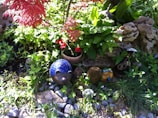A vibrant garden scene features a mix of lush green foliage and bright red flowers. Among the plants are decorative garden ornaments, including a blue hedgehog figurine and a whimsical frog figure. The setting includes natural rocks and pebbles, adding texture to the scene.