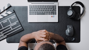 An overhead view featuring a person sitting at a desk with their hands clasped together, looking at a laptop on a dark mouse pad. To the left, there is a film clapperboard, and to the right, a pair of headphones and a computer mouse. The workspace is organized with tech and film elements.
