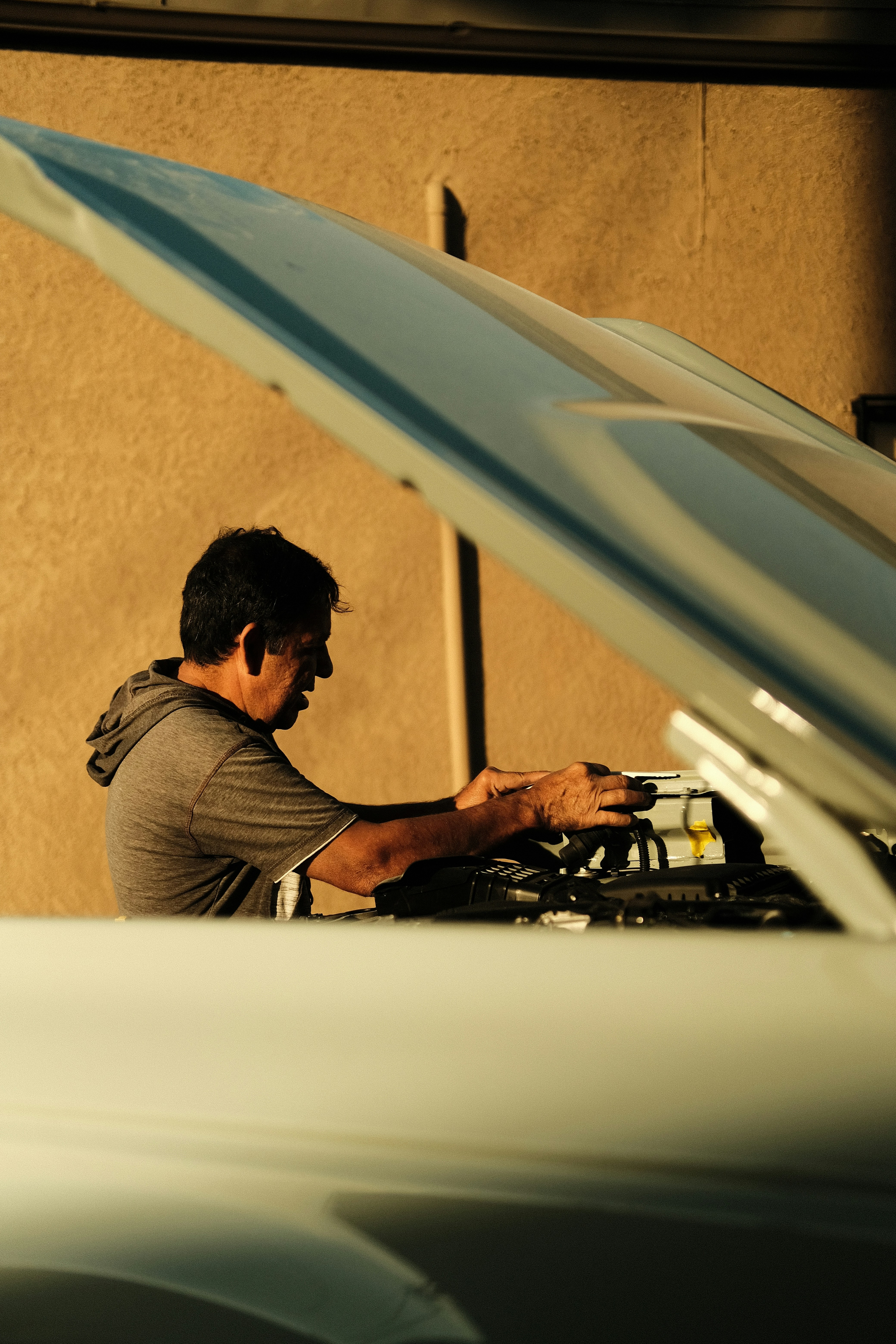 A man in a gray hoodie leans under the raised hood of a pale blue car, working on the engine in late sun against a stucco wall