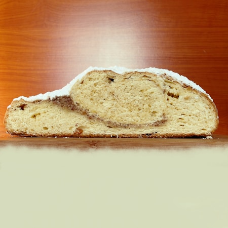 A cross-section of a loaf of bread with a crusty exterior and a swirled, textured interior. The surface is lightly dusted with a white powder, possibly flour or sugar. The background features a wooden surface and wall, enhancing the natural and rustic feel.