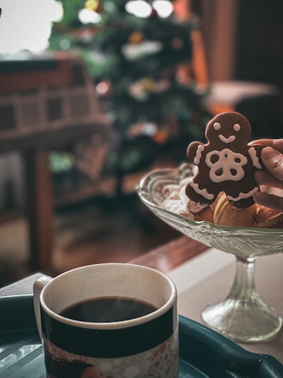 A gingerbread cookie decorated with white icing is held in a person's hand above a glass dish containing assorted baked goods. A mug filled with coffee is placed on a tray in the foreground. In the background, a blurred Christmas tree with colorful lights can be seen.