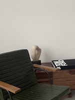 A sleek, modern interior setting with a dark green chair featuring wooden armrests in the foreground. A light-colored abstract sculpture sits on a wooden cabinet alongside a large book titled 'Tom Ford' with a black cover. The backdrop is a smooth, pale wall, providing a minimalist aesthetic.