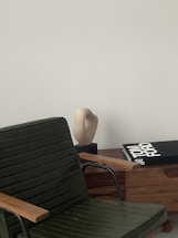 A sleek, modern interior setting with a dark green chair featuring wooden armrests in the foreground. A light-colored abstract sculpture sits on a wooden cabinet alongside a large book titled 'Tom Ford' with a black cover. The backdrop is a smooth, pale wall, providing a minimalist aesthetic.
