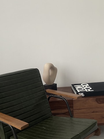 A sleek, modern interior setting with a dark green chair featuring wooden armrests in the foreground. A light-colored abstract sculpture sits on a wooden cabinet alongside a large book titled 'Tom Ford' with a black cover. The backdrop is a smooth, pale wall, providing a minimalist aesthetic.