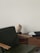 A sleek, modern interior setting with a dark green chair featuring wooden armrests in the foreground. A light-colored abstract sculpture sits on a wooden cabinet alongside a large book titled 'Tom Ford' with a black cover. The backdrop is a smooth, pale wall, providing a minimalist aesthetic.