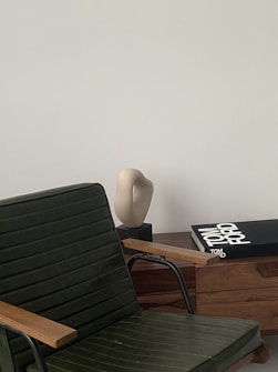A sleek, modern interior setting with a dark green chair featuring wooden armrests in the foreground. A light-colored abstract sculpture sits on a wooden cabinet alongside a large book titled 'Tom Ford' with a black cover. The backdrop is a smooth, pale wall, providing a minimalist aesthetic.