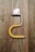 A piece of weathered wood with a mounted metal bracket in the shape of an upside-down 'U'. The upper part is a small, rectangular metallic hinge attached to the wooden surface with screws. A yellow, bent plastic or rubber tube is fitted onto the lower part of the metal bracket.