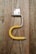 A piece of weathered wood with a mounted metal bracket in the shape of an upside-down 'U'. The upper part is a small, rectangular metallic hinge attached to the wooden surface with screws. A yellow, bent plastic or rubber tube is fitted onto the lower part of the metal bracket.