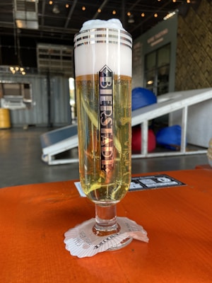 A tall glass filled with beer, topped with a thick layer of foamy head, placed on a vibrant orange table. The glass features a vertical logo with the name 'Bierstadt'. It sits on a coaster with printed text. In the background, there are elements of an industrial interior, including containers and blue and red cushions.