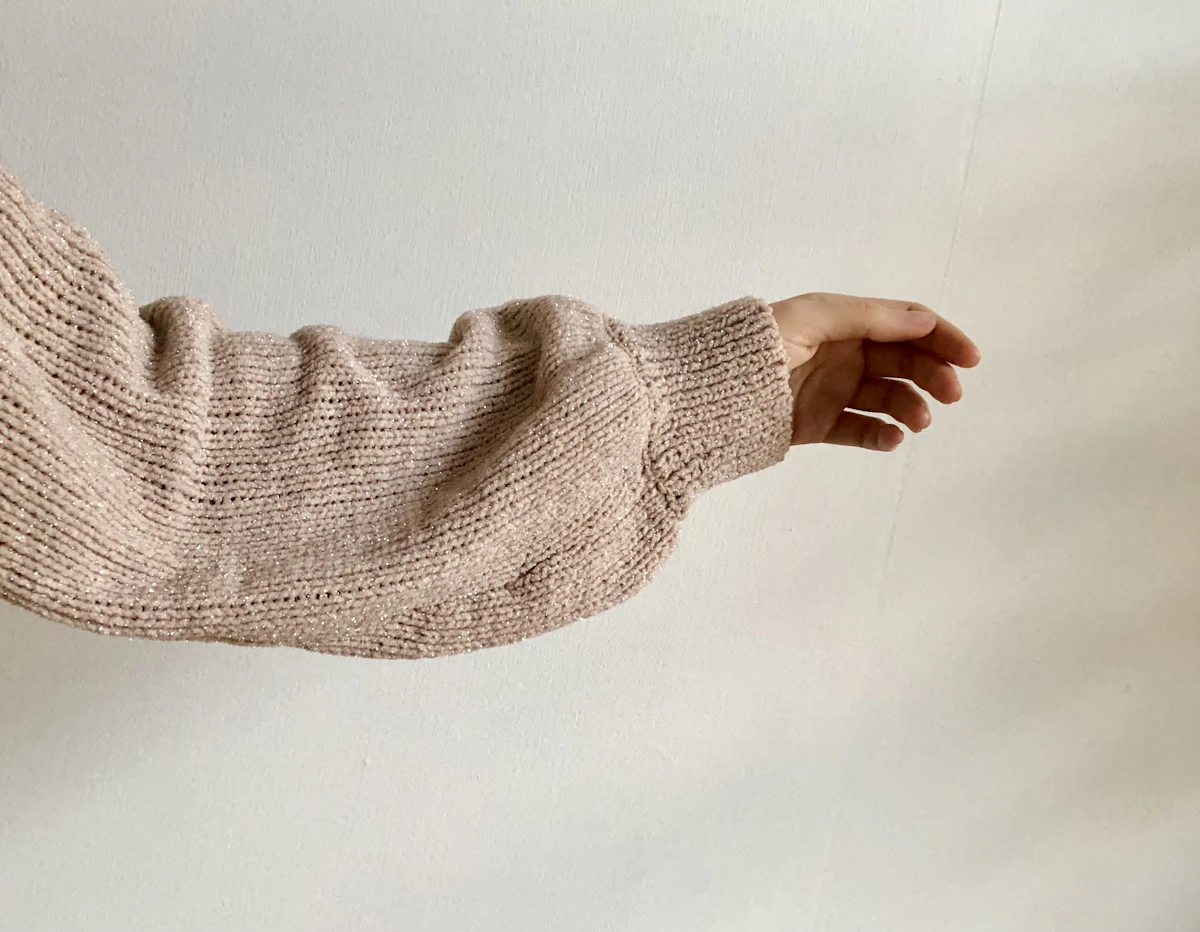 Lengan sweater rajut dengan tangan yang menjulur keluar melewati ujung lengan; pola rib pada kain terlihat jelas dan kainnya merenggang karena ditarik lengan.