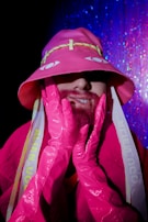 A person is wearing a bright pink outfit with long gloves and a matching hat. The hat has yellow accents and stylized eyes on it. The background features a shimmering blue and pink curtain, adding a party or disco atmosphere.