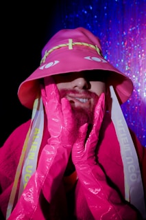 A person is wearing a bright pink outfit with long gloves and a matching hat. The hat has yellow accents and stylized eyes on it. The background features a shimmering blue and pink curtain, adding a party or disco atmosphere.