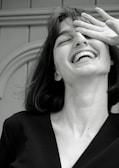 A monochrome photograph featuring a young woman with a joyful expression. Her head is tilted slightly back, her mouth open in laughter, and her eyes partially covered by her hand. The image captures a candid, carefree moment.