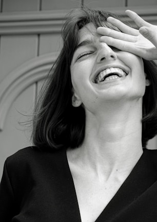 A monochrome photograph featuring a young woman with a joyful expression. Her head is tilted slightly back, her mouth open in laughter, and her eyes partially covered by her hand. The image captures a candid, carefree moment.
