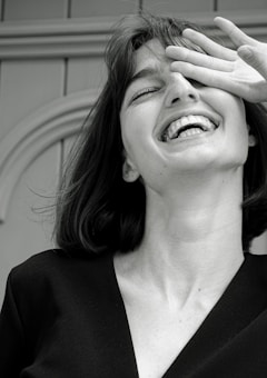 A monochrome photograph featuring a young woman with a joyful expression. Her head is tilted slightly back, her mouth open in laughter, and her eyes partially covered by her hand. The image captures a candid, carefree moment.